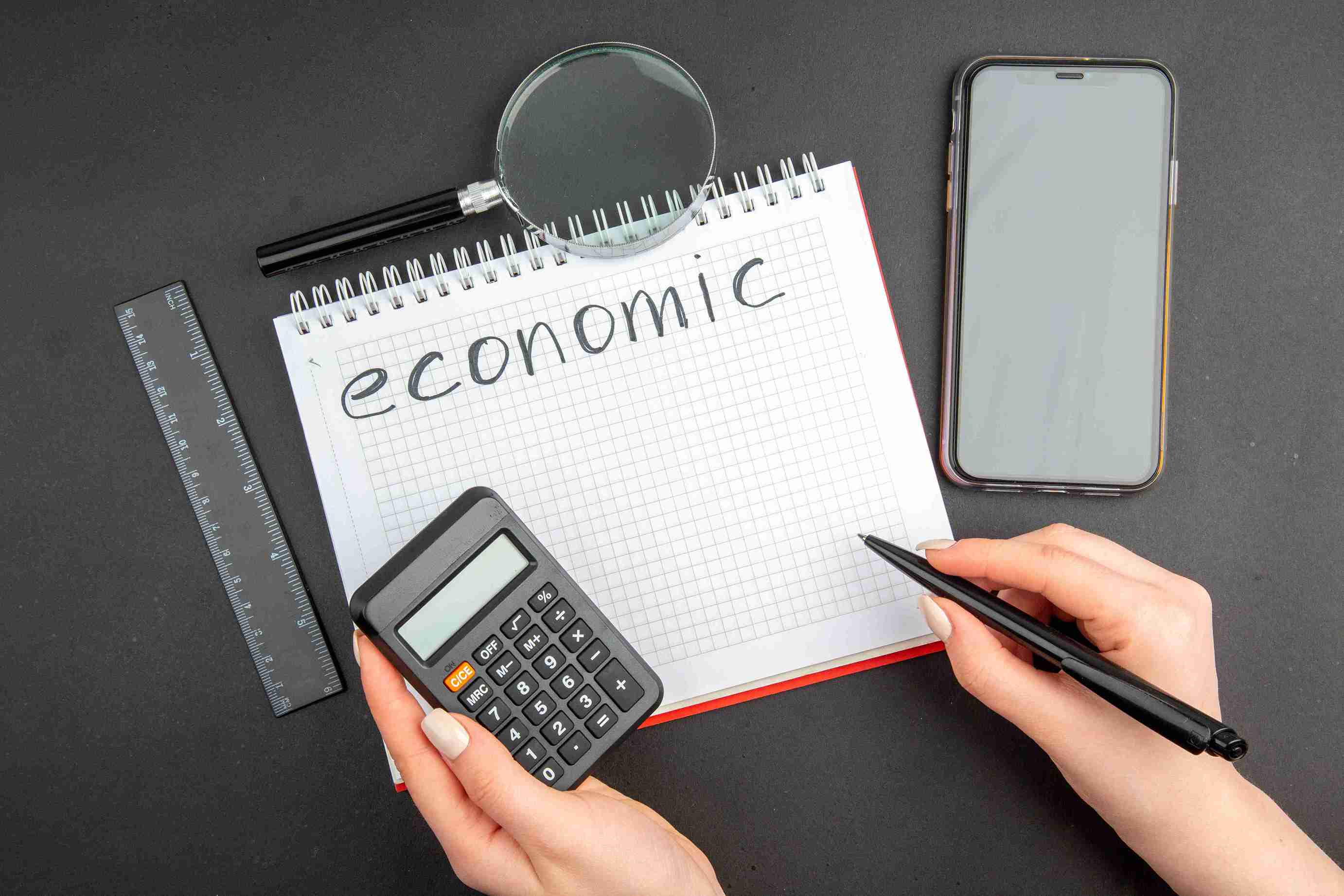 top-view-economic-written-spiral-notepad-lupa-phone-ruler-pen-calculator-female-hands-dark_11zon